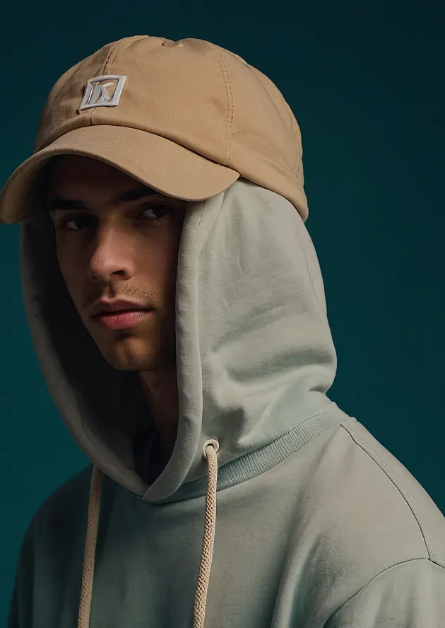 Young man wearing a tan baseball cap and a light green hoodie with the hood up against a dark teal background.
