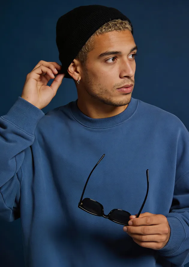 Young man with curly blond hair wearing a black beanie and blue sweatshirt holding black sunglasses, looking to the side against a dark blue background.